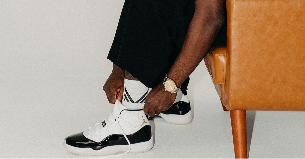 Man sitting on a brown leather chair wearing a black cap, black shirt, and white sneakers.